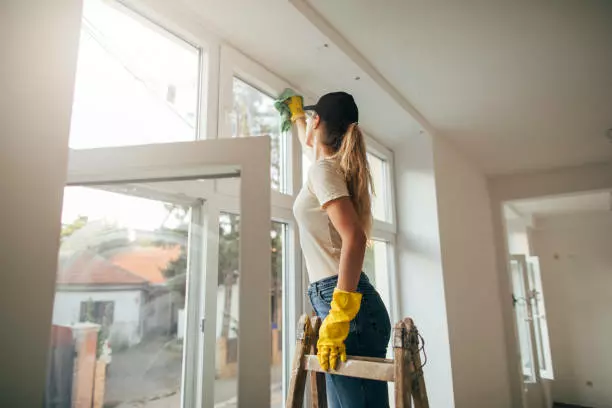 A woman providing maid service cleaning a window with a cloth while standing on a ladder indoors.