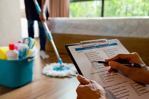 Person holding a deep cleaning checklist clipboard with another individual mopping the floor in the background.