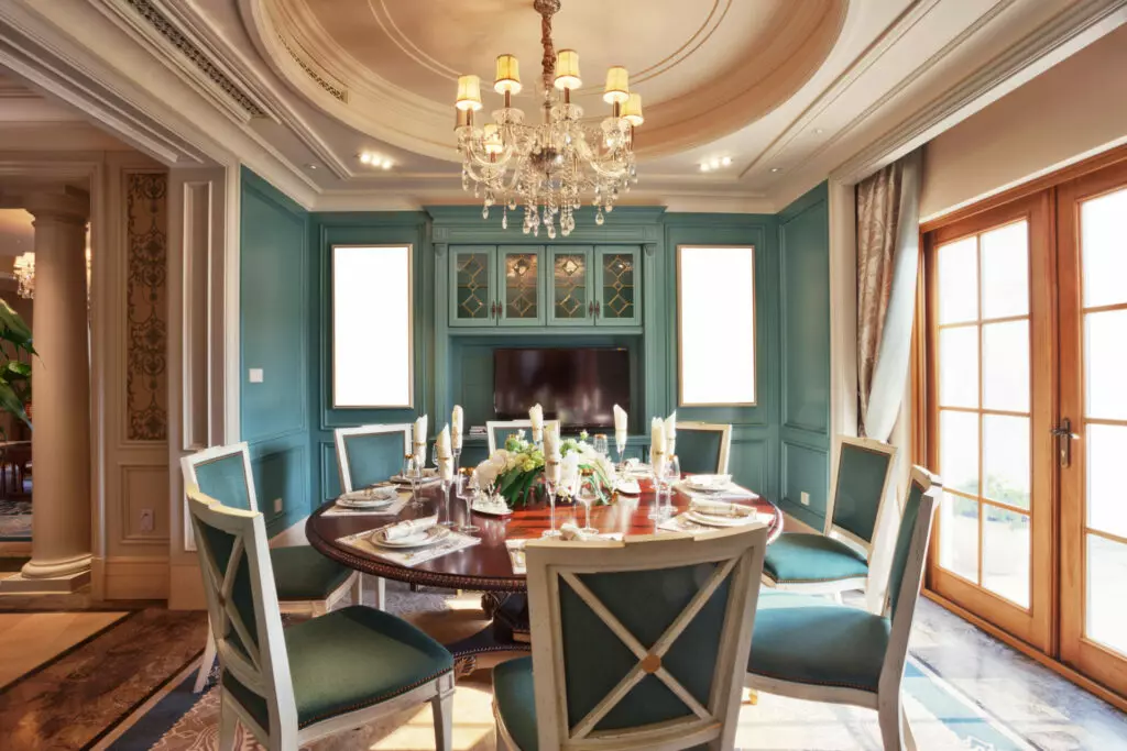 Elegant dining room with a round table set for a meal, featuring teal upholstered chairs, chandeliers, and French doors, maintained by an exceptional maid service.