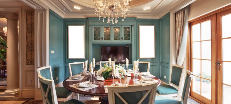 Elegant dining room with a round table set for a meal, featuring teal upholstered chairs, chandeliers, and French doors, maintained by an exceptional maid service.